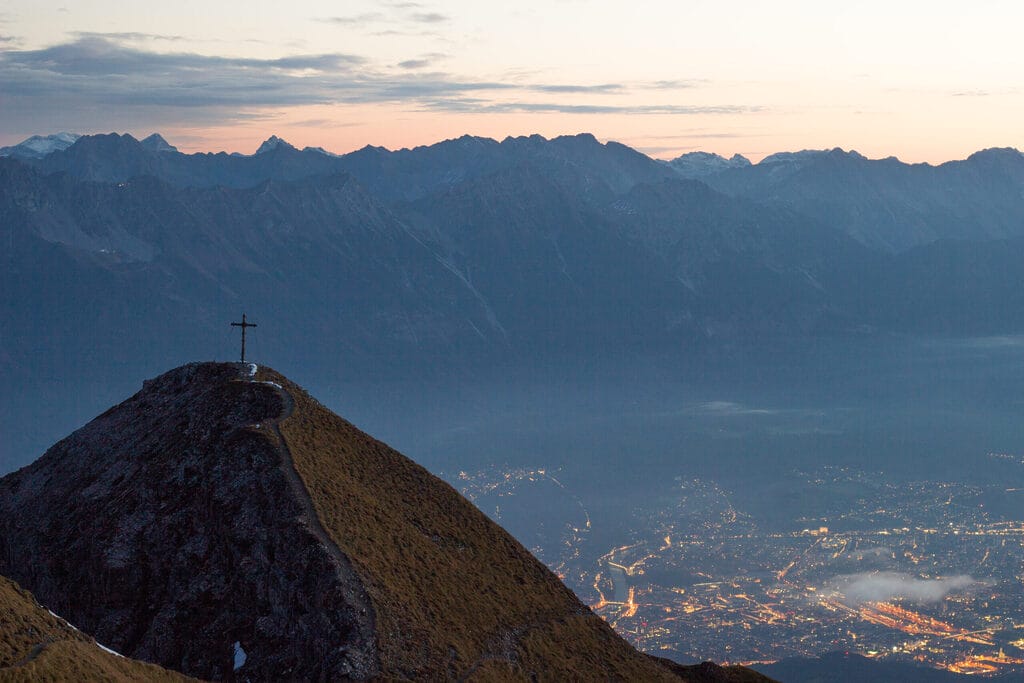 Auf dem Bild ist ein Gipfel mit einem Gipfelkreuz zu sehen. Im Hintergrund sieht man eine Bergkette und den Blick auf die Lichter einer Stadt. Die Stimmung ist mystisch und kurz vor Sonnenaufgang.
Die meisten deutschen wandern in die Schweiz aus.