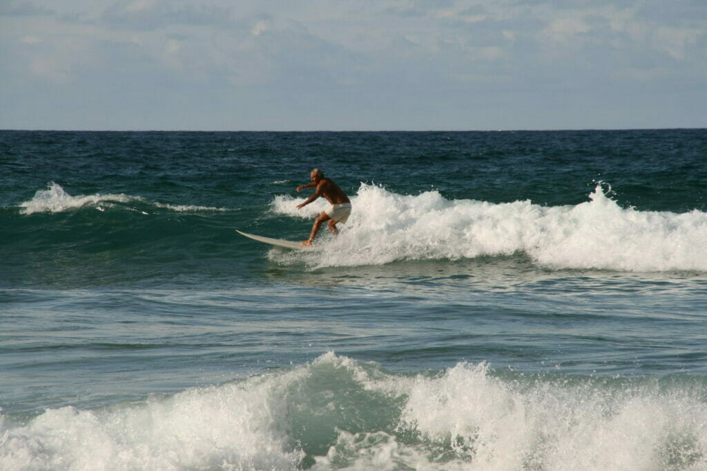 Dieses Foto zeigt einen älteren Herren in einer weißen Badehose beim Surfen in einer Welle.