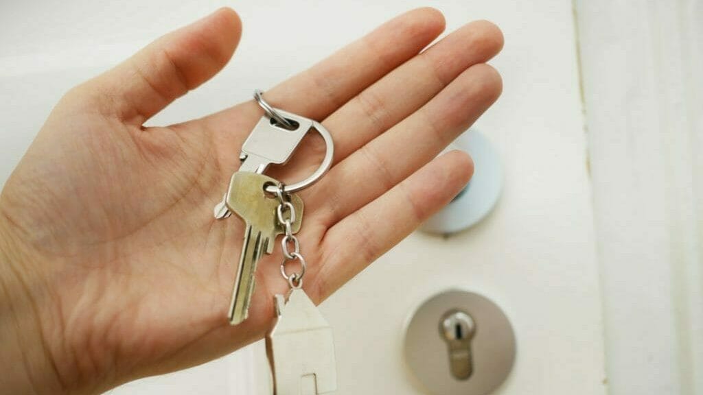 Eine Person hält einen Schlüssel in er Hand, der einen Anhänger, mit einem Haus daran hat. Im Hintergrund sieht man das Schlüsselloch einer Haus- oder Wohnungstüre.