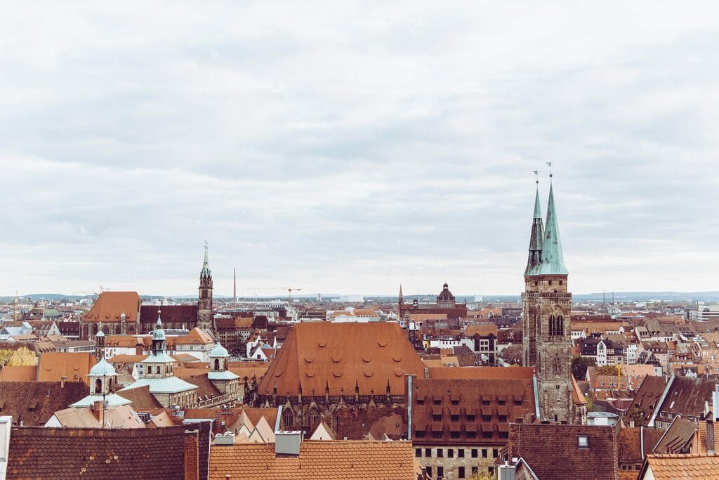 Auf dem Bild sieht man rote Dächer einer Stadt. Zudem sieht man drei Türme, die über die Dächer hinausragen, mit grünen Dächern.